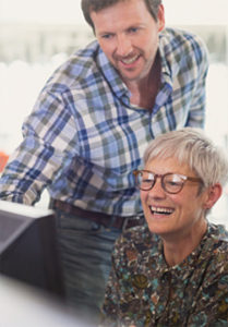 A woman sits smiling at a computer while a man stands behind her, showing her something on the screen.