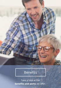 A woman sits smiling at a computer while a man stands behind her, showing her something on the screen. An option on the bottom of the image reads, "Benefits."