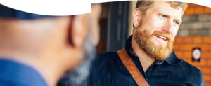 A bearded man stands talking to another man.