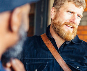 A bearded man stands talking to another man.