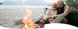 An older couple hold each other while sitting by a campfire on the beach.